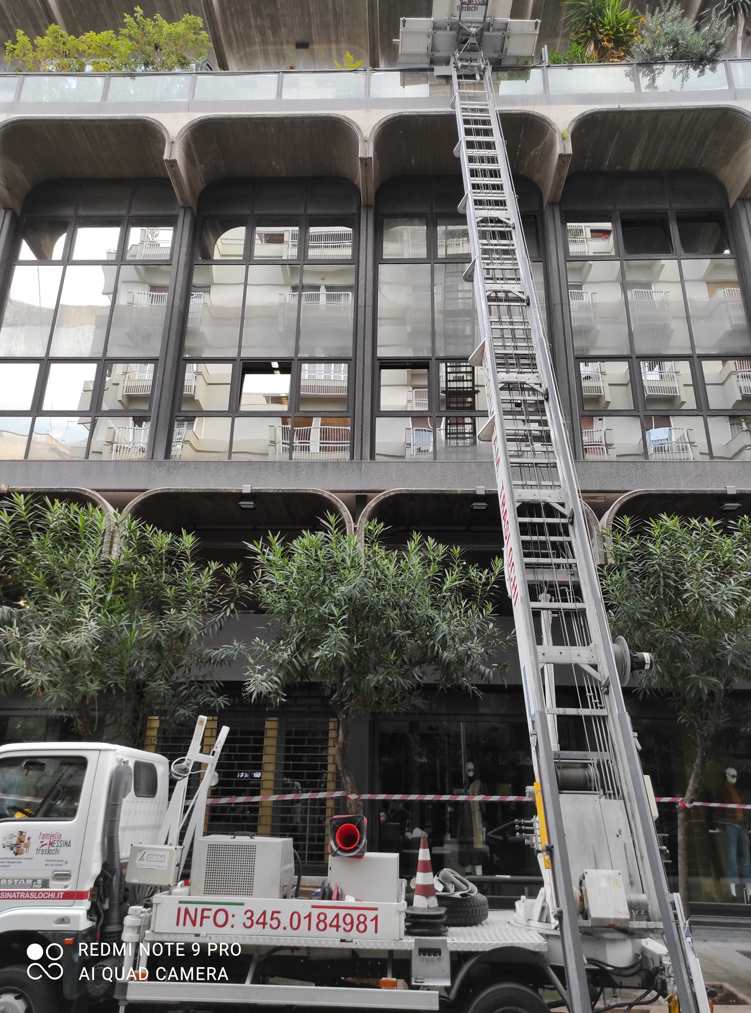 Scala montacarichi Famiglia Messina su camion con scritta INFO: 345.0184981 davanti a un palazzo con vetrate e oleandri