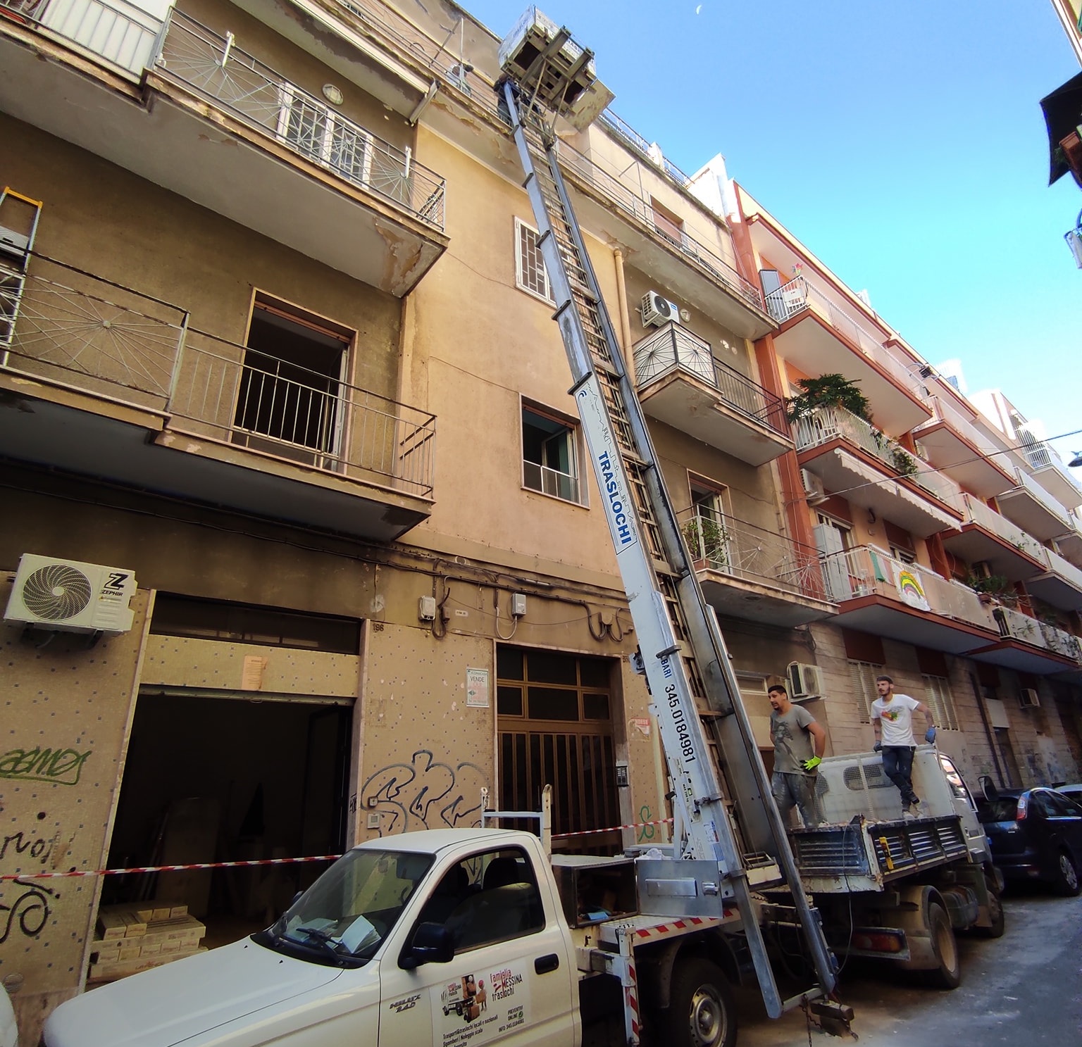 Autoscala Famiglia Messina Traslochi, numero 345 0184981, appoggiata a palazzo con balconi e due operai sul camion